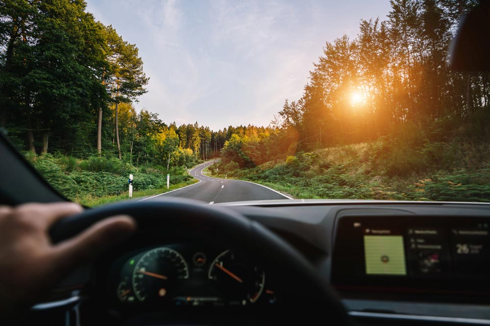 Händer på ratt i bil som kör på motorväg med skog och solnedgång framför vindrutan